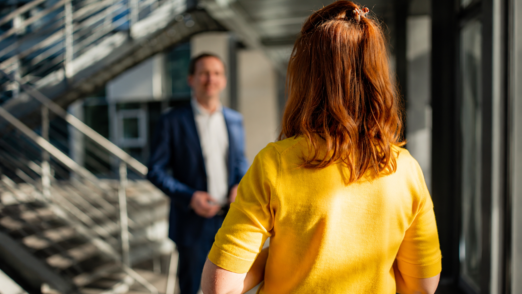 Frau mit rotem Haar in gelbem Shirt steht vor einem Mann in Anzug in einem modernen Gebäude.