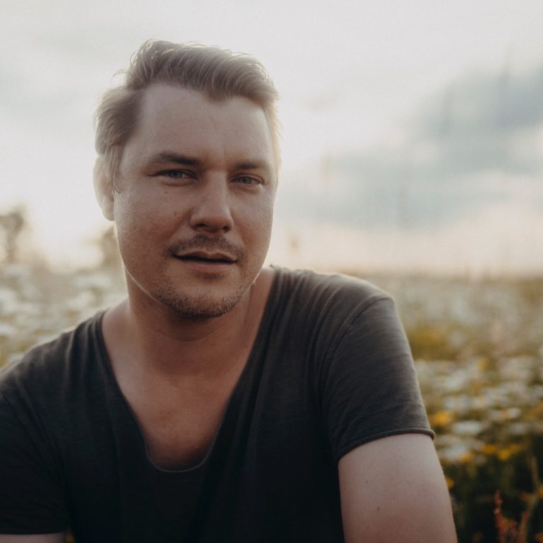 Junger Mann mit lässigem Oberteil, lächelt in einer Blumenwiese bei Sonnenuntergang.