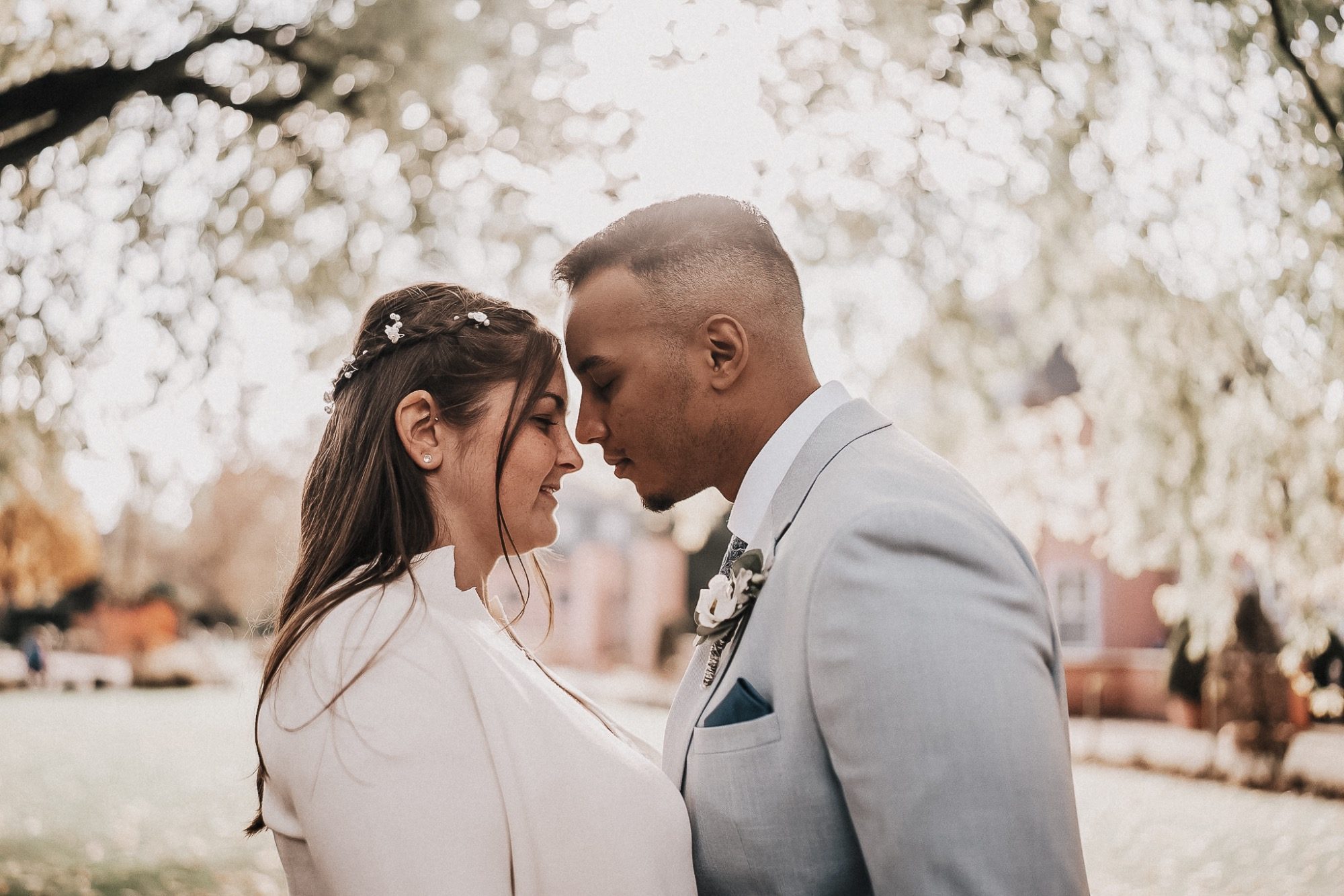 Braut und Bräutigam stehen sich gegenüber, ihre Stirnen berühren sich, umgeben von Bäumen.