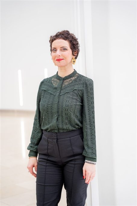 Frau mit lockigem Haar, grünem Oberteil und schwarzer Hose in einem hellen Raum.