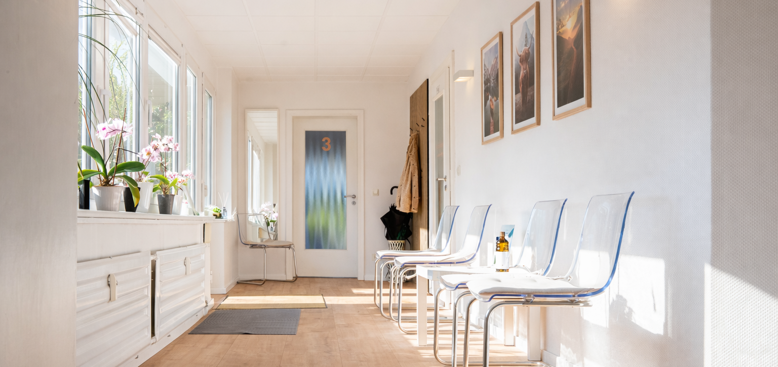 Helles Wartezimmer mit weißen Stühlen, Pflanzen und Bildern an der Wand.
