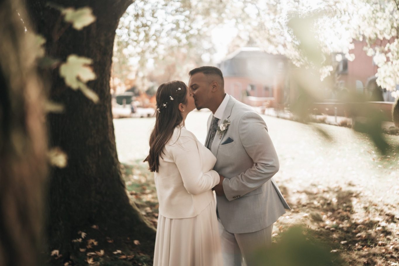 Ein Brautpaar küsst sich in einem sonnigen Park mit Bäumen im Hintergrund.